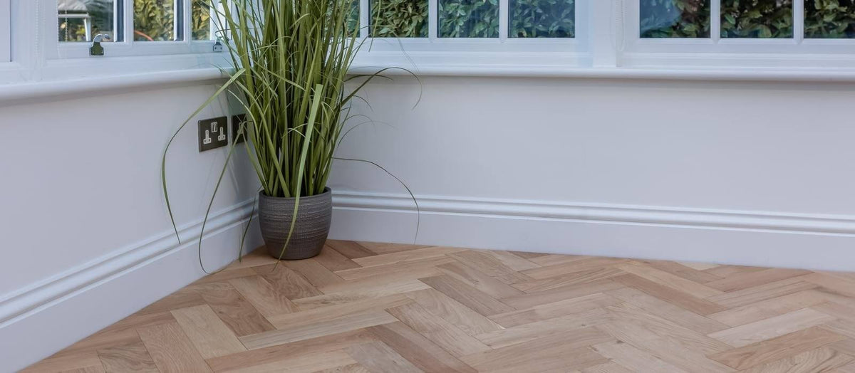 A corner of a room with a pot plant sitting on a light oak engineered herringbone parquet floor.
