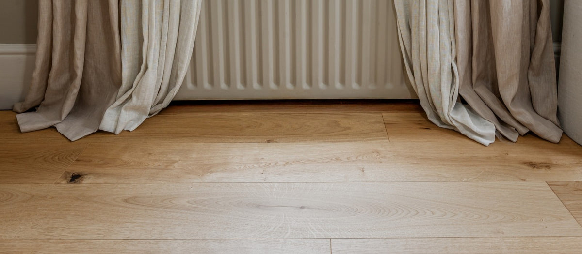 Light oak hardwood plank flooring with visible wood grain, styled in a room setting with beige curtains gathered around a white radiator.